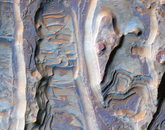 Multicolored ridges that appear to form cliffs and valleys. These layers, on the floor of an eroding impact crater, are extremely thin. Below the ridges are additional dark-toned deposits, exhibiting a variety of textures, some owing to the transport of material. The light-toned ridges are capped by thin dark-toned layers, and similar layers are exposed on the flanks. These layers are likely harder than the rest of the material and armor the surface against erosion by the wind.